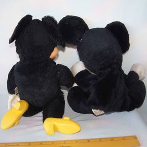 May include: Two black plush toys, one with a yellow shoe and the other with a white shoe, are sitting on a white surface. The toys are facing each other.