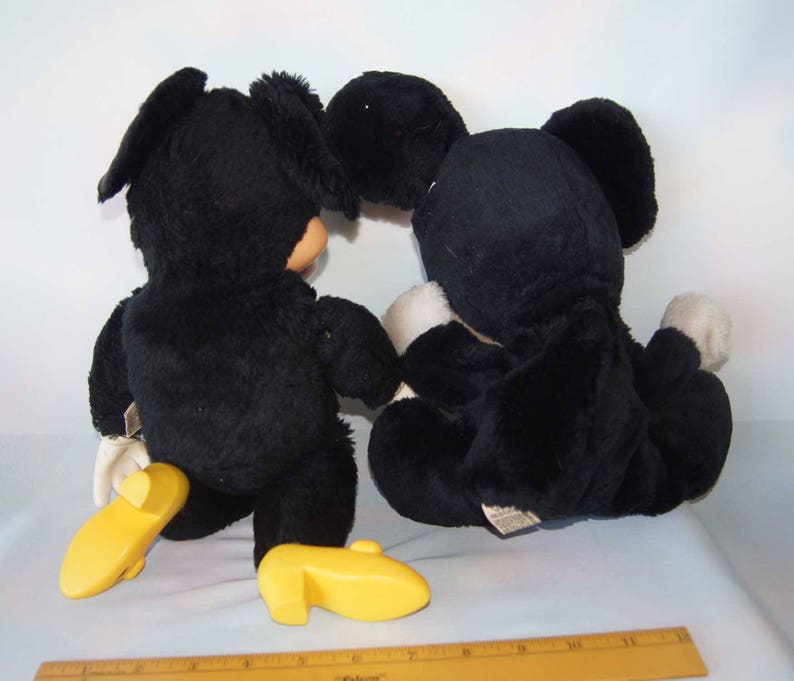 May include: Two black plush toys, one with a yellow shoe and the other with a white shoe, are sitting on a white surface. The toys are facing each other.