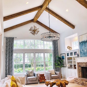 Op de afbeelding: Een woonkamer met een groot raam, een witte hoekbank, twee grijze fauteuils, een houten salontafel en een kroonluchter die hangt aan een gewelfd plafond met zichtbare houten balken.