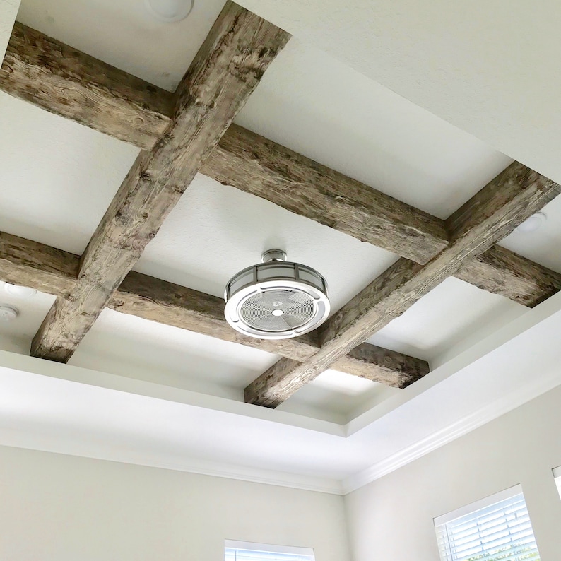 Op de afbeelding: Een wit plafond met een decoratief ontwerp van houten balken. Een plafondventilator met een zilveren afwerking is in het midden van de kamer geplaatst.