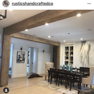 Op de afbeelding: Een grote, rustieke houten balk is ge&iuml;nstalleerd als decoratieve plafondbalk in een modern huis. De balk is in een lichte bruine kleur gebeitst en heeft een natuurlijke houtnerf. De balk is ge&iuml;nstalleerd boven een eettafel met stoelen en een witte marmeren open haard.