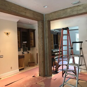 Op de afbeelding: Een kamer in aanbouw met zichtbare houten balken en een werknemer die een ladder gebruikt om het plafond te bereiken. De vloer is bedekt met roze afdekzeilen en er zijn twee ladders in de kamer.