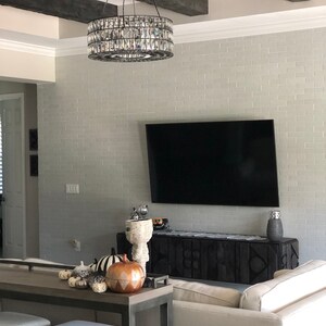 Op de afbeelding: Een woonkamer met een zwart-wit gepatroneerde bank, een zwart houten entertainmentcenter en een grote flatscreen-tv die aan een witte bakstenen muur is gemonteerd. Een kristallen kroonluchter hangt aan het plafond boven de tv. Op een lange houten tafel op de voorgrond staan decoratieve pompoenen.