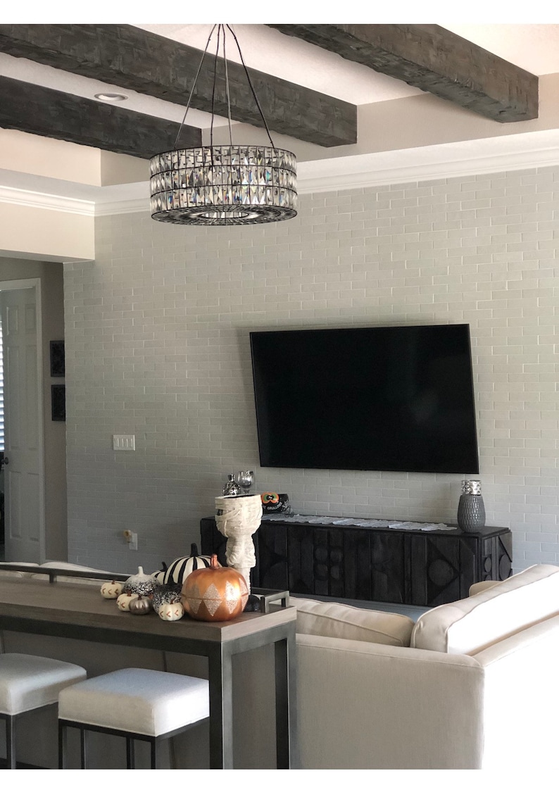 Op de afbeelding: Een woonkamer met een zwart-wit gepatroneerde bank, een zwart houten entertainmentcenter en een grote flatscreen-tv die aan een witte bakstenen muur is gemonteerd. Een kristallen kroonluchter hangt aan het plafond boven de tv. Op een lange houten tafel op de voorgrond staan decoratieve pompoenen.