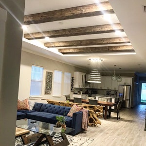Op de afbeelding: Een woonkamer met een blauwe hoekbank, een glazen salontafel en een wit tapijt. De kamer heeft een hoog plafond met zichtbare houten balken en een kroonluchter. De keuken is zichtbaar op de achtergrond.