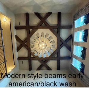 Op de afbeelding: Moderne houten balken in een kamer met een plafondventilator en ramen. De balken zijn donkerbruin gebeitst en hebben een zwarte wasbehandeling.