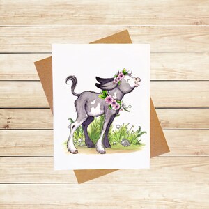 May include: A watercolor illustration of a gray and white donkey with pink flowers around its neck and head. The donkey is standing in a field of green grass with white flowers.