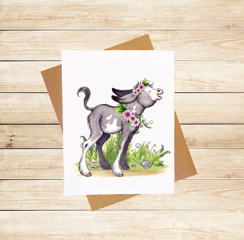 May include: A watercolor illustration of a gray and white donkey with pink flowers around its neck and head. The donkey is standing in a field of green grass with white flowers.