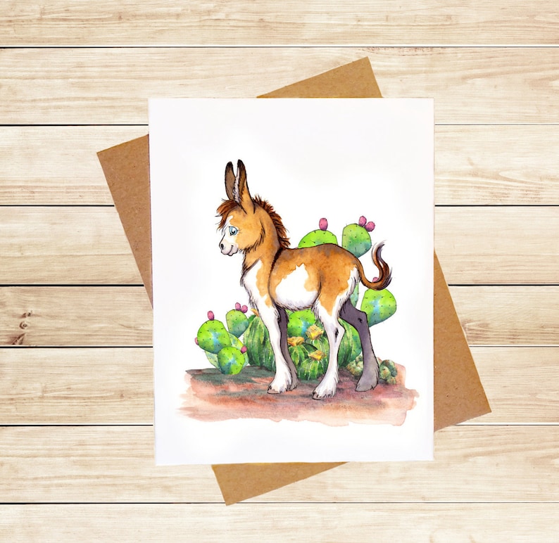 May include: A watercolor painting of a brown and white donkey standing in front of a prickly pear cactus. The donkey has blue eyes and is looking to the left. The cactus is green with pink flowers.
