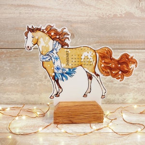 May include: A brown and white horse with a blue and white scarf stands on a wooden base. The horse is a January calendar with the days of the month printed on the base.