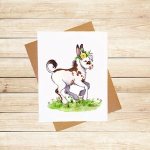 May include: A watercolor illustration of a white and brown spotted donkey with a yellow flower crown. The donkey is running through a field of green grass with white flowers.