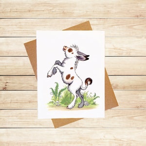 May include: A watercolor illustration of a white and brown spotted donkey with a gray head and hooves, standing on its hind legs with its front legs raised in the air. The donkey is surrounded by green grass and flowers.
