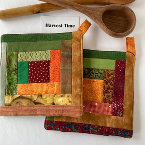 Puede incluir: Dos agarraderas acolchadas con un diseño de patchwork en tonos verdes, naranjas y marrones, con un borde marrón. El texto "Harvest Time" está en una tarjeta blanca. Dos cucharas de madera también están en la imagen.