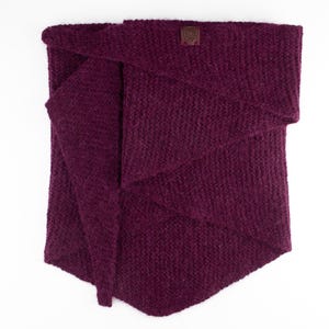 May include: A deep purple knitted cowl scarf with a textured, garter-stitch pattern. The scarf features an asymmetrical, triangular drape and includes a small, square brown leather tag stitched onto the upper center edge of the fabric.