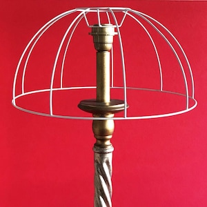 Puede incluir: Una lámpara de estilo vintage con una pantalla de alambre blanco. La lámpara tiene un portalámparas de color latón y una base decorativa. El tallo tiene un diseño retorcido plateado y dorado. El fondo es rojo.