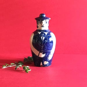 Op de afbeelding: Een blauw-witte keramische vaas in de vorm van een persoon met hoed en vest. De vaas heeft een strik en staat tegen een rode achtergrond. Een decoratief item.