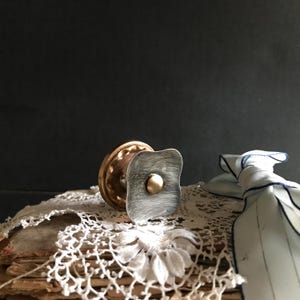 May include: A decorative cabinet knob with a square, brushed silver face and a small, round brass center. The knob is attached to a brass base. The knob rests on a stack of old books covered with white lace.