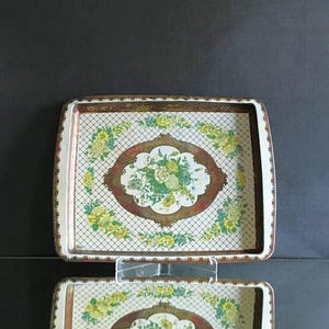 Puede incluir: Bandeja rectangular de metal con diseño floral. La bandeja presenta un fondo blanco con un patrón de cuadrícula, un óvalo central con flores y detalles florales amarillos. El borde es marrón con un borde festoneado.