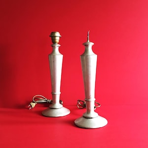 May include: Two off-white table lamps with a slender, elongated design. One lamp has a brass bulb socket, the other a decorative finial. Both lamps have a round base and are set against a vibrant red background.