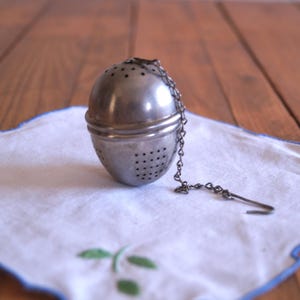 Op de afbeelding: Een zilverkleurige roestvrijstalen thee-ei in een bolvorm, voorzien van kleine gaatjes voor het trekken van thee. De infuser bevat een dunne metalen ketting met een klein haakje, waarmee het aan de rand van een kop of mok kan worden bevestigd.