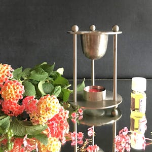 May include: A silver essential oil diffuser with a candle holder and a small bottle of oil. The diffuser has a round base and a curved top. Flowers with orange, yellow, and pink blooms are in the foreground.