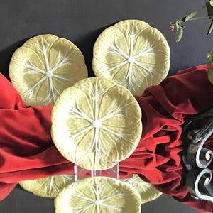 Puede incluir: Tres platos decorativos con diseño de hoja de col. Los platos son de color amarillo pálido con vetas blancas y se exhiben sobre una tela roja. Los platos son redondos y tienen un borde festoneado, y están sobre soportes transparentes.