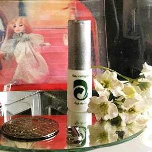 Puede incluir: Una botella de "Eau cologne" con tapón plateado y etiqueta blanca con logo verde y texto "Api-derm". La botella está sobre una superficie espejada con una moneda decorativa, un pequeño anillo plateado y flores blancas.