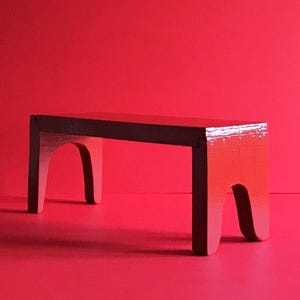 Puede incluir: Un pequeño banco de madera rojo con una parte superior marrón oscuro. El banco tiene cuatro patas curvas y está sobre un fondo rojo liso. El banco mide aproximadamente 76 cm de largo.