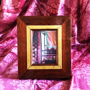 Op de afbeelding: Een rechthoekige houten fotolijst met een gouden rand. De lijst bevat een foto van een kamer met kleurrijke gordijnen en een donkere balustrade. De lijst staat tegen een roze bloemenachtergrond.