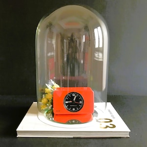 May include: A red alarm clock with black hands and numbers is displayed under a glass dome. The clock is sitting on a white book with the text "modern style 03" printed on the cover. A small bouquet of yellow flowers is visible next to the clock.