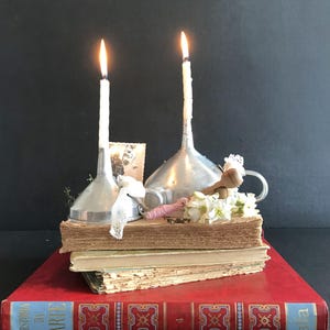 Op de afbeelding: Twee zilveren trechters met brandende kaarsen erop, gerangschikt op een stapel oude boeken met een vintage kantdoek en witte bloemen. Het onderste boek heeft de titel "Storia dell'Arte" op de rug gedrukt.