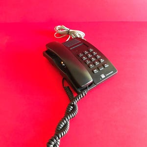 Op de afbeelding: Een zwarte bedrade vaste telefoon met een standaard numeriek toetsenbord met de cijfers nul tot en met negen, een asterisk, een hekje en een BIS-knop. Het apparaat bevat een zwart gekruld hoornsnoer en een grijze telefoonkabel.