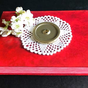 Può includere: Un oggetto in ottone è posto su un centrino di pizzo bianco sopra un libro rosso. Fiori bianchi sono disposti sulla copertina del libro. L'oggetto in ottone ha una forma circolare con un foro centrale. La copertina del libro ha un aspetto strutturato.