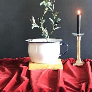 May include: A white enamel chamber pot with a dark blue rim and handle, containing a small artificial olive tree branch with green leaves and black olives. Beside it stands a tall brass candlestick holding a thin, lit black taper candle.