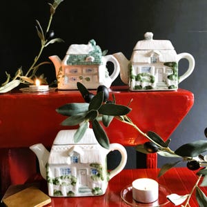 Può includere: Tre teiere in ceramica bianca a forma di case con finiture verdi e dettagli di edera. Le teiere sono appoggiate su un tavolo rosso con un ramo di ulivo verde e candele.