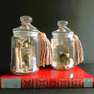 Può includere: Due barattoli di vetro trasparente con coperchi, riempiti di vari oggetti, tra cui una foto in bianco e nero, fiori secchi e altri oggetti effimeri. Ogni barattolo ha un fiocco di colore crema attaccato al coperchio.