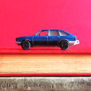 Può includere: Un'auto giocattolo blu e nera, con ruote nere e paraurti bianco. L'auto è appoggiata su una pila di libri con copertine rosse e beige, su uno sfondo rosso. Un giocattolo vintage.