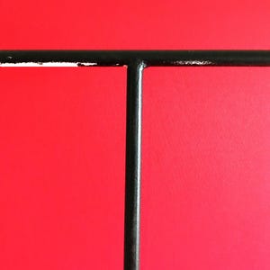 Puede incluir: Un objeto negro en forma de T de metal sobre un fondo rojo. La barra horizontal tiene algo de pintura blanca descascarillada. El objeto parece ser un expositor o un artículo similar.