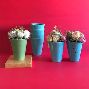 Puede incluir: Un conjunto de cuatro tazas cónicas de cerámica con acabado moteado. Una taza es de color verde claro, mientras que las otras tres son azul claro. Dos de las tazas azules contienen arreglos florales con rosas blancas y rosas.