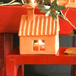 Puede incluir: Un portavelas de terracota con forma de casa con una vela pequeña en su interior. El portavelas está sobre una mesa de madera roja.
