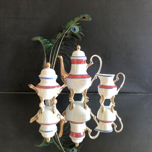 Pode incluir: Um conjunto de chá de porcelana branca com detalhes dourados e bordas vermelhas e azuis. O conjunto inclui uma chaleira, um cremeira e um açucareiro, todos sobre uma superfície reflexiva. Penas de pavão no fundo.