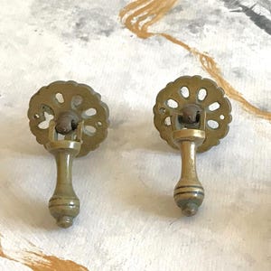 May include: Two brass drawer pulls featuring a circular backplate with a scalloped edge and a perforated floral pattern. Each pull has a hinged, vertical drop handle with a tapered, cylindrical shape and horizontal grooved detailing at the base.