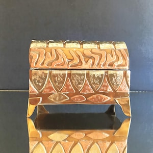 May include: A decorative metal box with a hinged lid. The box is primarily copper-colored with gold accents. The lid and sides feature intricate embossed patterns, including curved lines and triangular shapes. The box is elevated on four small feet.