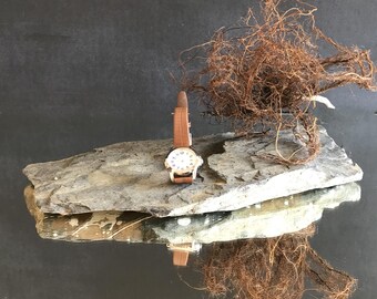Könnte beinhalten: Eine Vintage-Armbanduhr mit braunem Lederarmband und weißem Zifferblatt mit schwarzen Ziffern. Die Uhr liegt auf einer grauen Steinplatte, darüber eine Masse brauner Wurzeln. Die Szene spiegelt sich in einer dunklen, glänzenden Oberfläche.