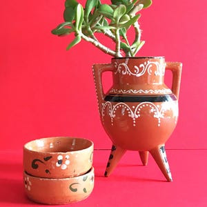 Puede incluir: Un jarrón de terracota de tres patas con dos asas, decorado con patrones pintados en blanco y negro, que contiene una planta suculenta verde. Junto a él hay dos cuencos de terracota apilados con motivos florales pintados a mano en blanco y negro.