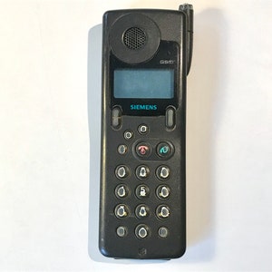 Puede incluir: Un teléfono móvil Siemens GSM negro con teclado y pantalla pequeña. El teléfono tiene un altavoz en la parte superior y una antena. El logotipo de Siemens es visible. El teléfono está sobre una superficie blanca.