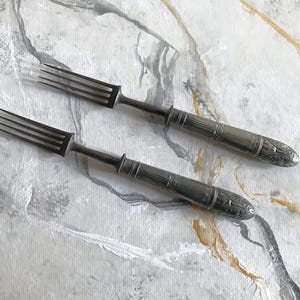Puede incluir: Dos tenedores de metal plateado con cuatro dientes y detalles en relieve ornamentados en los mangos cilíndricos. Los tenedores tienen un acabado pulido con patrones decorativos cerca de la base del mango, con un diseño esbelto.