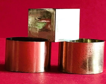 Può includere: Tre portacandele in ottone, uno quadrato e due rotondi, su uno sfondo rosso.