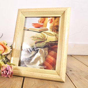 May include: A light wood picture frame with a white mat and a printed image of a room interior. The image shows a book, a bowl, and a pillow with a floral pattern. The text "Osterley Collection" is visible at the bottom of the image.