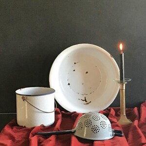 Puede incluir: Un cuenco de esmalte blanco con manchas de óxido, un cubo de esmalte blanco con asa de alambre y borde azul, un colador de esmalte gris claro con mango largo y un candelabro de latón con una vela negra encendida sobre tela roja.